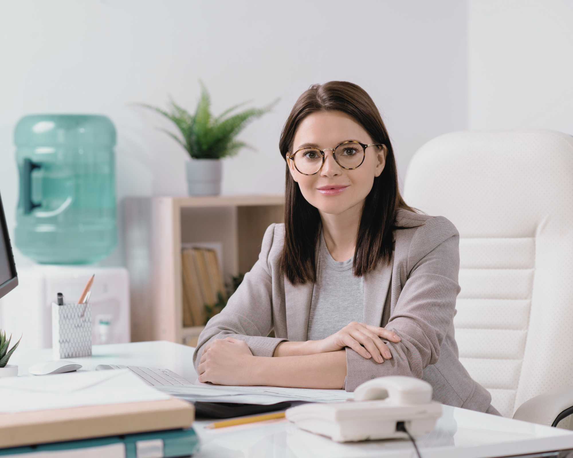 Person mit Brille sitzt an Büroschreibtisch mit Laptop und Telefon und blickt lächelnd in die Kamera