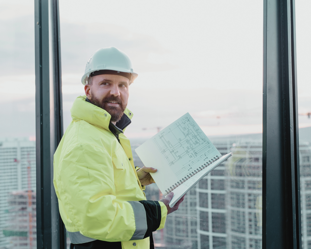 Person in Arbeitsschutzkleidung Jacke und Helm steht an einem großen Fenster und überblick eine Großbaustelle