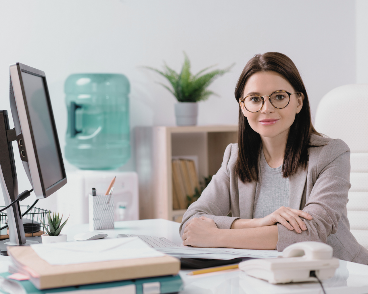 Frau mit Brille sitzt selbstbewusst an einem Büroschreibtisch mit Computer und Unterlagen